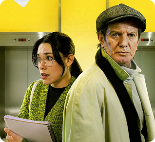 Una mujer con gafas sostiene unas carpetas y permanece nerviosa junto a un hombre mayor con gorra y abrigo, frente a dos puertas de ascensor y una pared de color amarillo brillante.