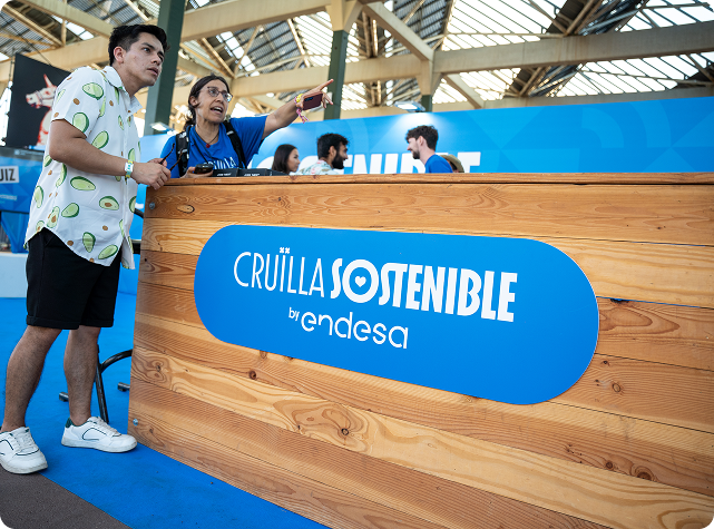 Un joven y una mujer junto a una caseta de madera con un cartel azul en el que se lee "CRUÏLLA SOSTENIBLE by Endesa". La mujer señala hacia delante, y el fondo muestra a otras personas en un espacio abierto y cubierto.