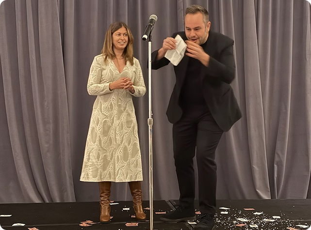 Una mujer con un vestido blanco estampado está de pie junto a un hombre vestido de negro, que realiza un truco de magia con una servilleta en el escenario. Hay cartas y trozos de papel esparcidos por el suelo. Un micrófono se interpone entre ellos.